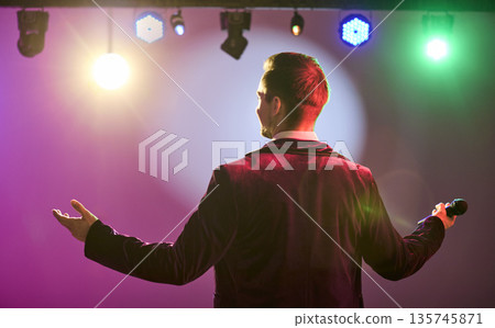 Showman with microphone on stage under colorful lights, back view, open arms Showman with microphone on stage under colorful lights, back view, open arms 135745871