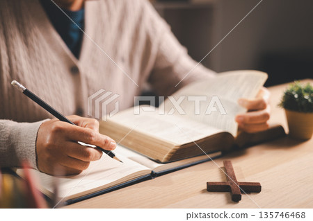 Person taking notes while reading an open Bible at a desk, pencil and journal beside a wooden cross, warm indoor light and soft focus, conveying Christian faith, devotion and scripture study at home. Person taking notes while reading an open Bible at a desk, pencil and journal beside a wooden cross, warm indoor light and soft focus, conveying Christian faith, devotion and scripture study at home. 135746468