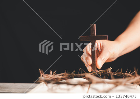 Hand placing a wooden cross above a crown of thorns on an open Bible, dark background and dramatic light with copy space, symbolizing crucifixion, sacrifice, Holy Week and Easter faith. Hand placing a wooden cross above a crown of thorns on an open Bible, dark background and dramatic light with copy space, symbolizing crucifixion, sacrifice, Holy Week and Easter faith. 135746475