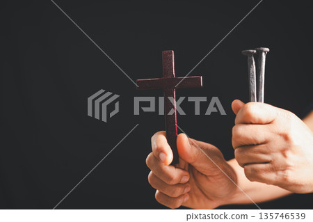 Hands holding a wooden cross and two iron nails on dark background, dramatic light and copy space, symbolizing crucifixion, sacrifice, Good Friday, Holy Week and Easter Christian faith. 135746539