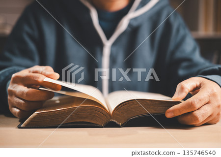 Man reading an open Bible at a desk, turning pages in warm indoor light with shallow depth of field, symbolizing Christian faith, devotion, study and quiet reflection at home. 135746540