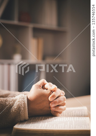 Clasped hands in prayer over an open book, warm light and shallow depth of field, symbolizing faith, devotion, reflection and hope for spiritual guidance during quiet study at home. 135746554