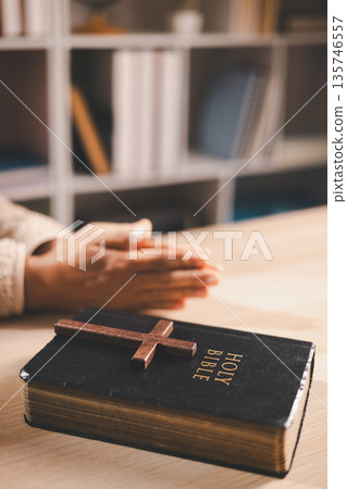 Wooden cross resting on a Holy Bible on a table with blurred praying hands in warm indoor light, symbolizing Christian faith, devotion, worship and quiet reflection at home with copy space for text. Wooden cross resting on a Holy Bible on a table with blurred praying hands in warm indoor light, symbolizing Christian faith, devotion, worship and quiet reflection at home with copy space for text. 135746557