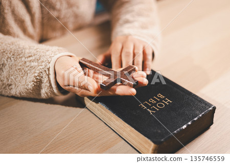 Hands holding a wooden cross above a Holy Bible on a table, warm indoor light and soft focus, symbolizing Christian faith, prayer, devotion and hope during quiet worship at home with copy space. Hands holding a wooden cross above a Holy Bible on a table, warm indoor light and soft focus, symbolizing Christian faith, prayer, devotion and hope during quiet worship at home with copy space. 135746559