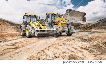 Construction site shows dry, cracked earth with dirt mounds. Two loaders work on the ground while clouds cover part of the sky Construction site shows dry, cracked earth with dirt mounds. Two loaders work on the ground while clouds cover part of the sky 135746589