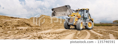 A wheel loader moves across a sandy area, leaving deep tracks. A large pile of sand is visible in the background, indicating ongoing construction work A wheel loader moves across a sandy area, leaving deep tracks. A large pile of sand is visible in the background, indicating ongoing construction work 135746591