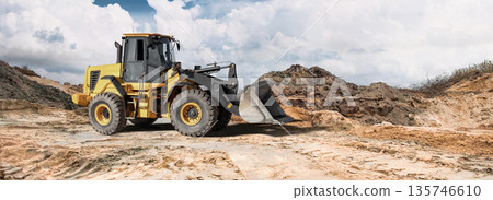 Loader operates on dry, cracked ground with dirt mounds. Partly cloudy sky adds contrast to the construction activity in the area 135746610