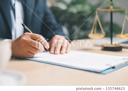 Lawyer in suit signing a contract on clipboard at office desk with justice scales in background, representing legal services, compliance review, agreement approval and documentation. 135746623