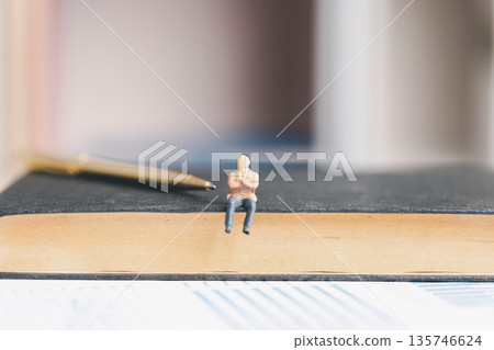 Miniature business figure sitting on a thick book with pen and blurred office background, symbolizing strategy, leadership, decision making, planning and corporate finance analysis. 135746624