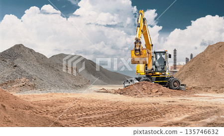A wheeled excavator works at a construction site, moving sand for construction work, surrounded by piles of building materials 135746632