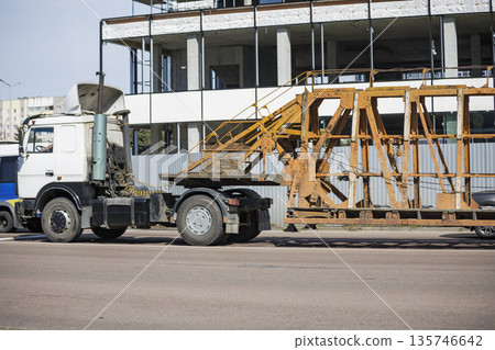 A truck drives on a road, transporting a metal ramp, while construction workers are present nearby at a building site 135746642