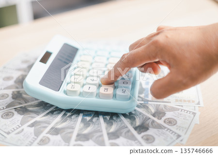 Hand pressing a pastel calculator on scattered US dollar bills, calculating budget, expenses, savings and cash flow for personal finance, investment planning and cost control at home or office. Hand pressing a pastel calculator on scattered US dollar bills, calculating budget, expenses, savings and cash flow for personal finance, investment planning and cost control at home or office. 135746666