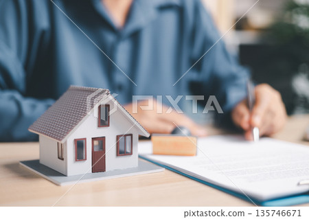 Person signing a real estate contract on clipboard beside a house model and keys, closing a home purchase, mortgage or rental agreement, property insurance and loan approval at office desk. Person signing a real estate contract on clipboard beside a house model and keys, closing a home purchase, mortgage or rental agreement, property insurance and loan approval at office desk. 135746671