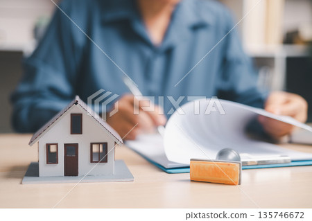 Person signing a real estate contract on clipboard beside a house model and keys, closing a home purchase, mortgage or rental agreement, property insurance and loan approval at office desk. Person signing a real estate contract on clipboard beside a house model and keys, closing a home purchase, mortgage or rental agreement, property insurance and loan approval at office desk. 135746672