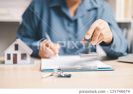 Person signing a real estate contract on clipboard beside a house model and keys, closing a home purchase, mortgage or rental agreement, property insurance and loan approval at office desk. Person signing a real estate contract on clipboard beside a house model and keys, closing a home purchase, mortgage or rental agreement, property insurance and loan approval at office desk. 135746674