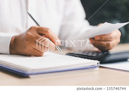 Accountant writing notes while checking a bill and using a calculator on desk, managing budgeting, bookkeeping, tax and expenses for financial planning, business accounting and audit. 135746744