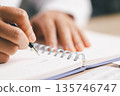 Business person taking notes in a spiral notebook beside a laptop and financial charts, planning strategy, budgeting and reporting for corporate analysis, meeting prep and productivity. 135746747