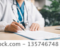 Doctor in white coat with stethoscope signing medical paperwork on clipboard at clinic desk, completing patient records, insurance forms, prescription approval and healthcare documentation. 135746748