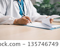 Doctor in white coat with stethoscope reviewing a report and signing medical documents on clipboard at clinic desk, handling patient records, referrals and insurance billing paperwork. 135746749