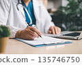 Doctor with stethoscope signing patient forms on clipboard beside laptop at clinic desk, completing electronic records, telemedicine notes, insurance paperwork and care documentation. 135746750