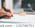 Close-up of hand holding pen over a document on clipboard with laptop blurred in background, signing agreement, reviewing paperwork for business approval and administration workflow tasks. 135746751