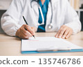 Doctor in white coat with stethoscope signing an approved medical form on clipboard at desk, representing healthcare documentation, prescription, insurance claims and patient care. 135746786