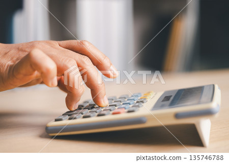 Close-up hand pressing a calculator on a wooden desk in an office, calculating costs for budgeting, accounting, tax, payroll and financial planning in a modern workplace, soft light. Close-up hand pressing a calculator on a wooden desk in an office, calculating costs for budgeting, accounting, tax, payroll and financial planning in a modern workplace, soft light. 135746788