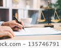 Lawyer in suit signing a contract on clipboard at office desk with justice scales in background, representing legal services, compliance review, agreement approval and documentation. 135746791