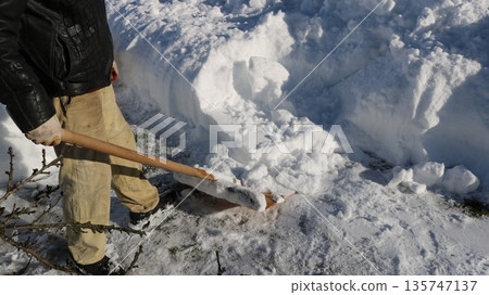 A partially visible worker with a wooden shovel standing near a large snowdrift on a sunny day, manually clearing snow from a site, pushing snow away with a shovel, assessing the work done on snow  135747137