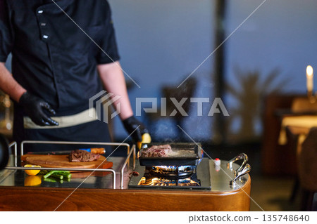 Chef Grills Meat On Portable Stove In Modern Restaurant Kitchen With Fresh Produce 135748640
