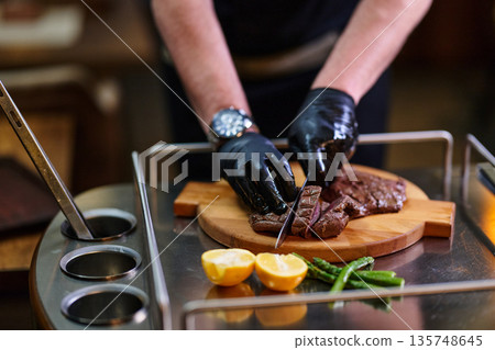 Chef Slicing Juicy Beef Steak On Cutting Board With Lemons And Green Beans In Kitchen Chef Slicing Juicy Beef Steak On Cutting Board With Lemons And Green Beans In Kitchen 135748645