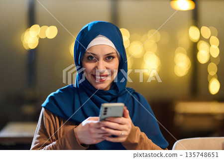 Smiling Woman In Blue Hijab Using Smartphone In Cozy Indoor Cafe Setting Smiling Woman In Blue Hijab Using Smartphone In Cozy Indoor Cafe Setting 135748651