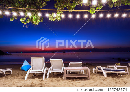 Relaxing beachfront lounge at night with white chairs, decorative string lights, and a vibrant blue and orange twilight sky, Koh Samed Relaxing beachfront lounge at night with white chairs, decorative string lights, and a vibrant blue and orange twilight sky, Koh Samed 135748774