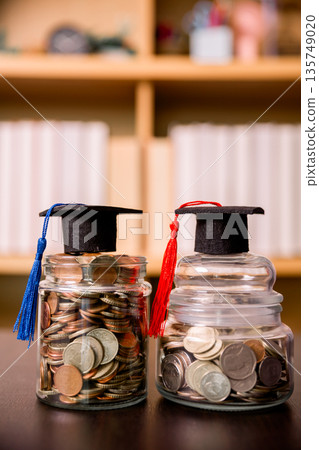 Two coin jars topped with graduation caps and tassels on a desk, suggesting education savings, tuition planning, scholarship funding and student budgeting with copy space. Soft library background. 135749020
