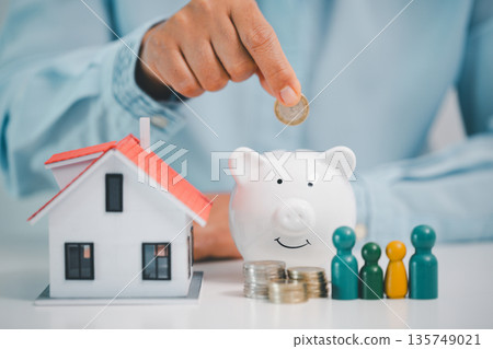 Hand dropping coin into piggy bank beside model house and family figures, symbolizing home savings, mortgage planning, downpayment budgeting and secure household finances for future homeowners. Hand dropping coin into piggy bank beside model house and family figures, symbolizing home savings, mortgage planning, downpayment budgeting and secure household finances for future homeowners. 135749021