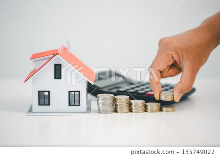 Hand stacking coins beside model house and calculator on white background, illustrating mortgage budgeting, home loan planning, down payment savings and real estate investment goals. Hand stacking coins beside model house and calculator on white background, illustrating mortgage budgeting, home loan planning, down payment savings and real estate investment goals. 135749022