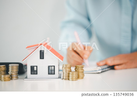 Model house with stacked coins on desk while person writes in notebook, illustrating home budget planning, mortgage payments, down payment savings and real estate goals with copy space. 135749036