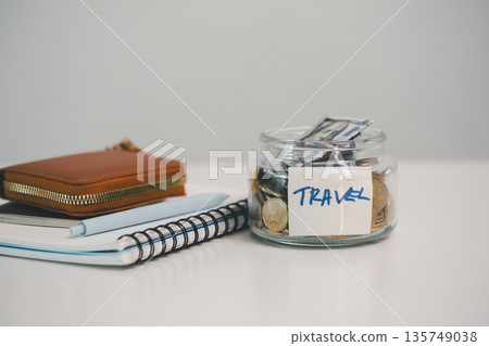 Glass jar labeled TRAVEL packed with coins and banknotes beside notebook, pen and wallet on white background, representing vacation savings, trip budgeting and expense tracking goals. 135749038