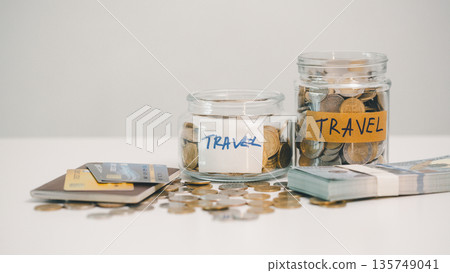 Two travel savings jars filled with coins beside credit cards, scattered change and a stack of cash on white background, symbolizing vacation fund goals, budgeting and smart spending. Two travel savings jars filled with coins beside credit cards, scattered change and a stack of cash on white background, symbolizing vacation fund goals, budgeting and smart spending. 135749041