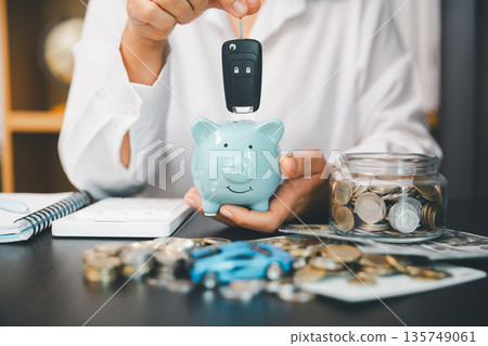 Hand holding car key above blue piggy bank beside coin jar, cash and calculator, showing car savings, auto loan planning, down payment budgeting and personal finance goals. 135749061