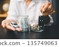 Hand holding a car key fob and miniature car beside jars of cash and coins, representing saving for a vehicle, down payment goals, auto loan planning and budgeting at home with copy space. 135749063