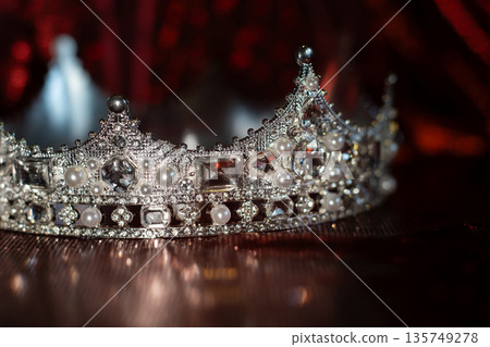 White crown with crystals, diamonds on dark red sparkle background. 135749278