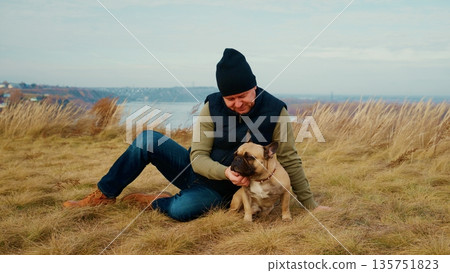 A Calm Moment in Nature: A Person Enjoys Quality Time with Their Adorable Dog on a Beautiful Scenic Landscape Surrounded by Tall Grass and Tranquil Views of the Water 135751823