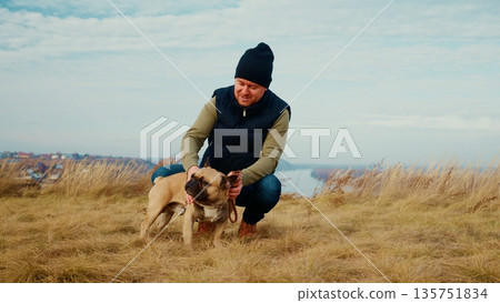 A Man Comforts His Dog While Enjoying Nature's Beauty on a Scenic Hilltop Overlooking a Serene Landscape Under a Clear Sky with Rolling Grasslands and Calm Waters 135751834