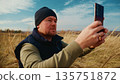 A Man in a Beanie Capturing a Photo with His Smartphone Amidst Tall Grass Under a Clear Blue Sky, Enjoying Nature and the Outdoors in a Peaceful Setting 135751872