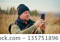 Man in Field Using Smartphone to Capture Nature's Beauty, Surrounded by Tall Grasses and Scenic Landscape Under Clear Sky, Enjoying Outdoor Photography Experience and Connection 135751896