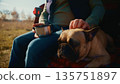 A Person Enjoys a Tranquil Moment Outdoors with Their Beloved Dog, Sitting on a Blanket While Petting the Animal, Surrounded by Nature Under a Clear Sky in a Serene Setting 135751897