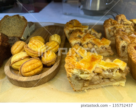 Fresh baked apple pie slices with golden crust are displayed alongside small sandwich cookies and bread on wooden boards Fresh baked apple pie slices with golden crust are displayed alongside small sandwich cookies and bread on wooden boards 135752437