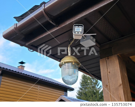 Outdoor security camera and light fixture mounted on house eave. Home surveillance system for safety and property protection. 135753074