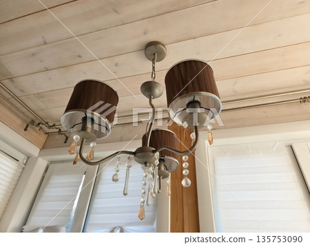 Chandelier with four lamps and decorative crystals hangs from a white wooden ceiling in a home living space with bright windows. Chandelier with four lamps and decorative crystals hangs from a white wooden ceiling in a home living space with bright windows. 135753090